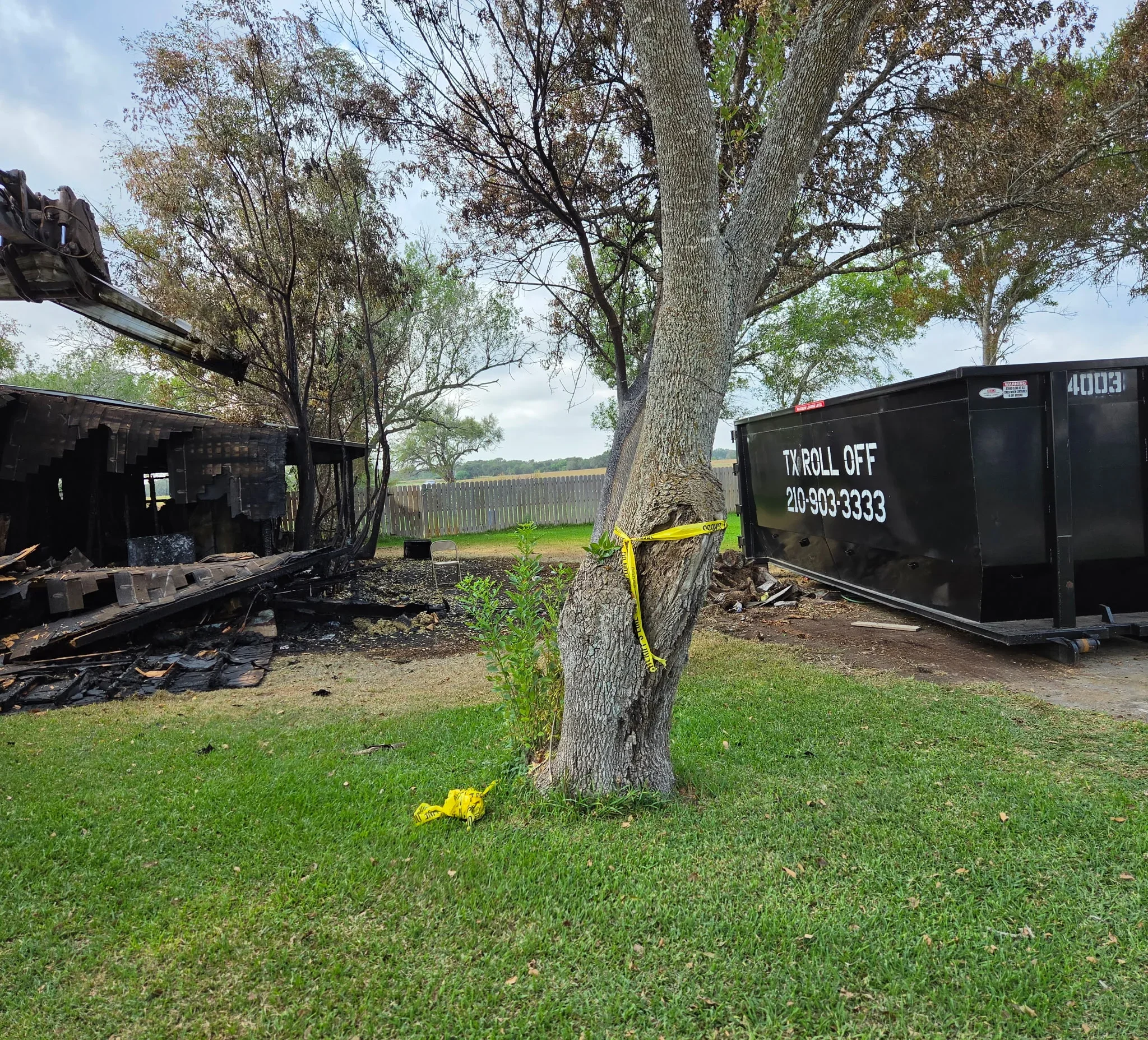 Black roll-off dumpster with yellow safety straps on residential lawn near large oak tree